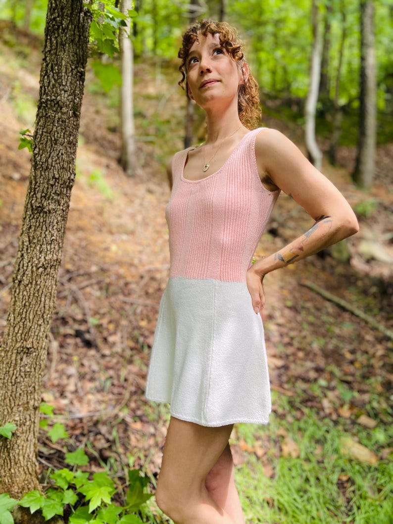 May include: A pink and white knitted dress with a sleeveless bodice and a flared skirt. The dress is worn by a person standing in a forest.