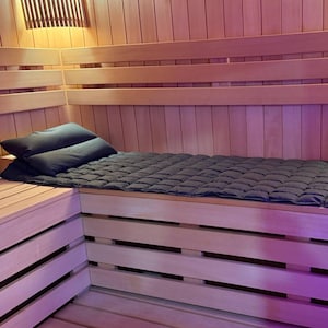 May include: Interior of a wooden sauna with a bench, pillows, and a quilted mat. The walls and ceiling are made of light-colored wood. Two dark pillows rest on the bench, and a dark quilted mat covers the seating area. The sauna is illuminated by warm lighting.