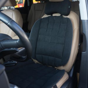 May include: Black car seat cushion and headrest set. The seat cushion has a quilted pattern and is placed on a tan car seat. A black headrest pillow is attached to the headrest. The car's steering wheel is visible.