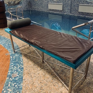 May include: A massage table with a brown cover and a teal-colored border. The table has a cylindrical bolster pillow and is supported by stainless steel legs. The background features a swimming pool and nautical-themed wall art.