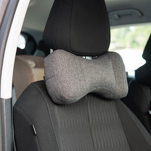 May include: A gray car seat headrest with a gray neck pillow attached. The pillow is shaped like a bow tie and has a textured fabric.