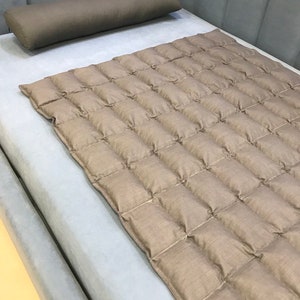 Op de afbeelding: Een bed met een grijze hoofdbord en een lichtgrijze matras. Een bruin rolkussen ligt op het bed. Een bruine, gewatteerde deken bedekt het bed en voegt textuur en warmte toe.