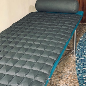 May include: A gray quilted massage table with a matching bolster pillow. The table has a teal blue trim and is supported by silver metal legs. The quilted surface is made of square padded sections.