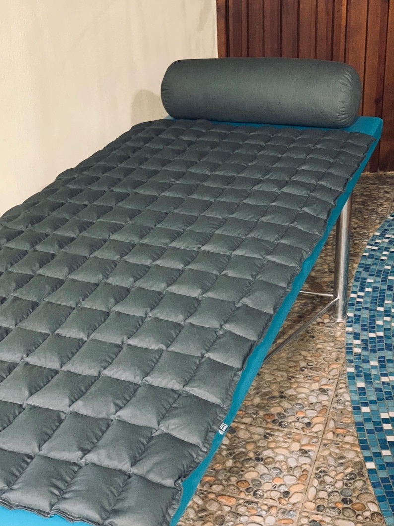 May include: A gray quilted massage table with a matching bolster pillow. The table has a teal blue trim and is supported by silver metal legs. The quilted surface is made of square padded sections.