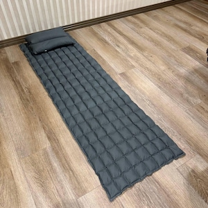 May include: A dark gray sleeping pad and pillow set. The pad is rectangular with a quilted pattern, and the pillow is a small, rectangular shape. The set is designed for camping or travel and is placed on a wooden floor.