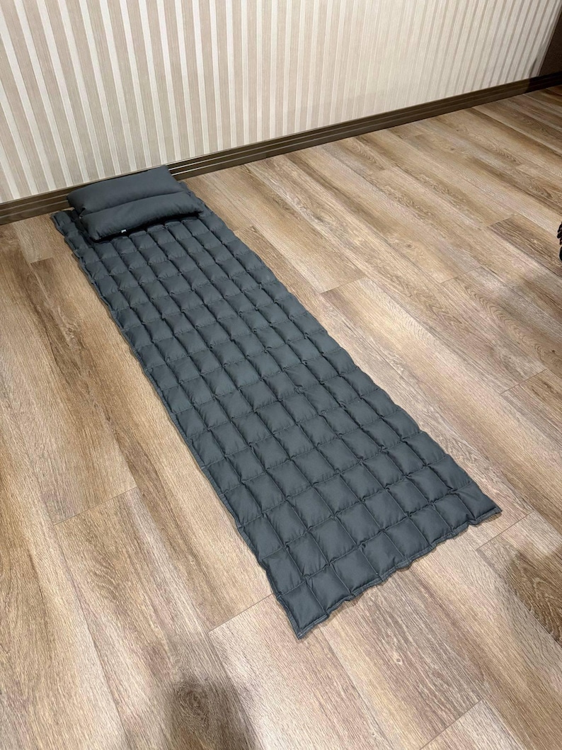 May include: A dark gray sleeping pad and pillow set. The pad is rectangular with a quilted pattern, and the pillow is a small, rectangular shape. The set is designed for camping or travel and is placed on a wooden floor.