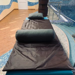 May include: A massage table with a dark brown cover and teal base is set up in a spa-like room. Two dark green pillows, one rectangular and one cylindrical, rest on the table. The room has a pool with a mural and mosaic tile accents.