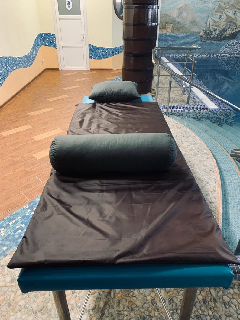 May include: A massage table with a dark brown cover and teal base is set up in a spa-like room. Two dark green pillows, one rectangular and one cylindrical, rest on the table. The room has a pool with a mural and mosaic tile accents.