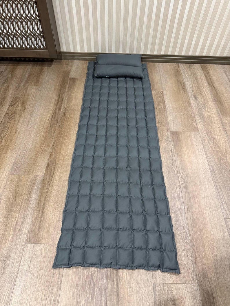 May include: A gray quilted sleeping mat with an attached pillow. The mat is rectangular with a grid pattern, and the pillow is a small, rectangular shape. The mat and pillow are made of a soft, padded material.