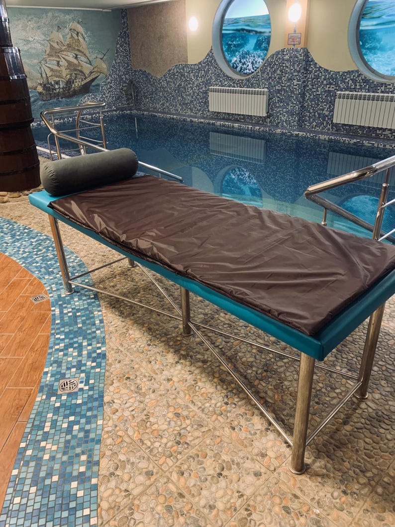 May include: A massage table with a brown cover and a teal-colored border. The table has a cylindrical bolster pillow and is supported by stainless steel legs. The background features a swimming pool and nautical-themed wall art.