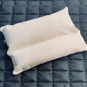 May include: A beige reading pillow with a rectangular shape and a built-in armrest. The pillow is made of a textured fabric and is designed for comfort and support while reading or relaxing. The pillow is placed on a dark blue quilted surface.