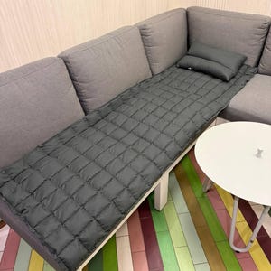 May include: A gray quilted mattress pad and pillow set on a gray sectional sofa. The pad is rectangular with a diamond pattern. A small white side table is next to the sofa. The floor has a colorful striped pattern.