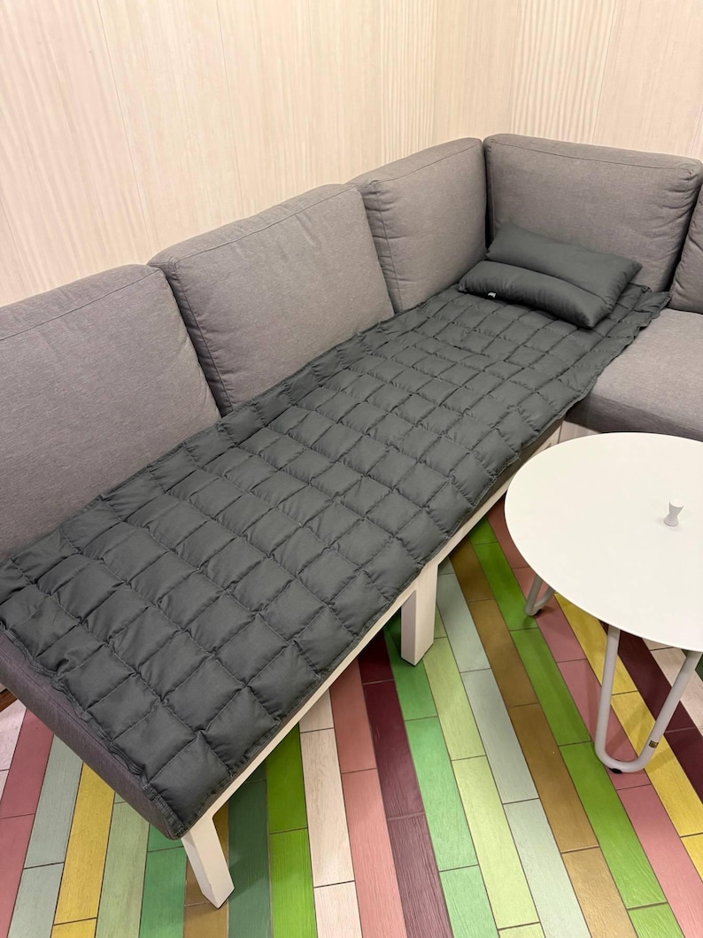 May include: A gray quilted mattress pad and pillow set on a gray sectional sofa. The pad is rectangular with a diamond pattern. A small white side table is next to the sofa. The floor has a colorful striped pattern.