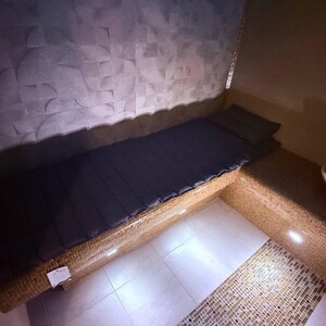 May include: A built-in bench with a dark blue mattress and pillow. The bench is made of woven material and is set against a textured gray wall. The floor is tiled with white squares. The scene is illuminated by recessed lighting.