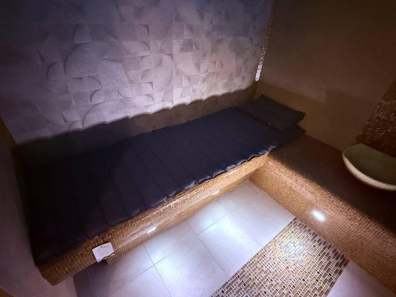 May include: A built-in bench with a dark blue mattress and pillow. The bench is made of woven material and is set against a textured gray wall. The floor is tiled with white squares. The scene is illuminated by recessed lighting.