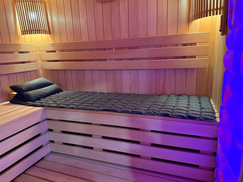 May include: Interior of a wooden sauna with a bench, pillows, and a quilted mat. The walls and ceiling are made of light-colored wood. Two dark pillows rest on the bench, and a dark quilted mat covers the seating area. The sauna is illuminated by warm lighting.