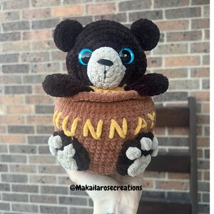 May include: A brown crocheted bear with blue eyes is sitting inside a brown crocheted pot with the word "HUNNY" written in yellow yarn.