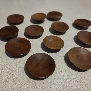 May include: A collection of twelve small, circular wooden knobs with a smooth, dark brown finish and visible wood grain patterns. Each knob features a slightly concave top surface and a rounded edge, designed for use as cabinet hardware.