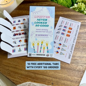 May include: A set of three rectangular paper pull tab game cards with light blue and white backgrounds. The center card features the text LUCKY 42 NEVER LOOKED SO GOOD, Stella's Birthday Souvenir Pull Tab, 10 WINNERS, and 08082026.