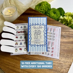 May include: A rectangular wedding pull tab game card with blue vertical stripes and a central illustration of a bride and groom. The card features the text Marv & Stella's Wedding, August 8th, 2026, and 21 Winners. It includes perforated tabs with various wedding-themed icons and prize designations.