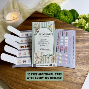 May include: A baby shower pull tab game card featuring a light blue and white striped design with illustrations of a hot air balloon, an elephant, and a gift box. The text reads Baby Smith's, Already stealing hearts and making us feel lucky.