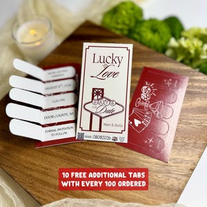 May include: A rectangular white and maroon save the date card featuring the text Lucky in Love and a casino-themed graphic. The card includes perforated pull-tabs with event details like August 8th, 2026, 5:00 PM, and Door County, WI.