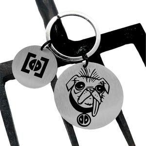 May include: Silver key chain with two round metal tags. One tag has a black and white graphic of a pug with its tongue sticking out. The other tag has a black and white graphic of a square with a circle inside.