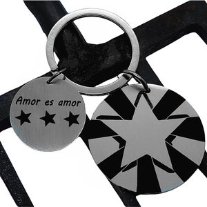 May include: A silver keychain with two round metal tags. One tag has the text "Amor es amor" with three stars. The other tag has a black and white starburst design with a silver star in the center.