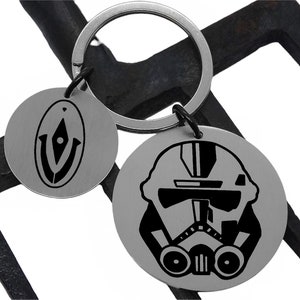 May include: A silver keychain with two round metal charms. One charm features a black and white graphic of a Star Wars Stormtrooper helmet. The other charm features a black and white graphic of a symbol.