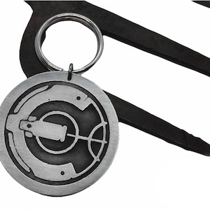 May include: A silver-colored metal keychain with a circular pendant featuring an engraved design. The design includes a central cylindrical shape with a crosshair-like element and other geometric details. The keychain is attached to a metal ring.