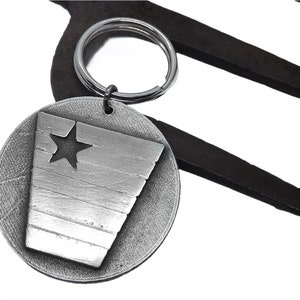 May include: A silver-colored keychain with a round tag featuring a cut-out star and a stylized flag design. The keychain is attached to a metal ring and set against a dark, abstract background.