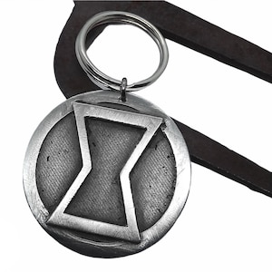 May include: A circular metal keychain featuring a raised hourglass symbol in the center. The piece has a brushed silver finish with a textured, dark gray background within the circle. A standard silver-toned split ring is attached at the top.