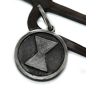 May include: A round, silver-colored keychain pendant with a raised hourglass symbol inside a circle. The pendant is attached to a silver keyring. The background includes a dark, textured metal object.