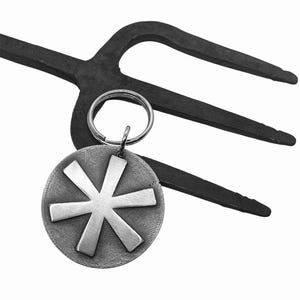 May include: A black metal garden fork with a silver-colored key ring and a round pendant. The pendant features a starburst design. The background is white.
