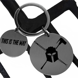 May include: A silver keyring with two round metal tags. One tag has the text "THIS IS THE WAY" and the other tag has a black and white illustration of a helmet with crossed swords.