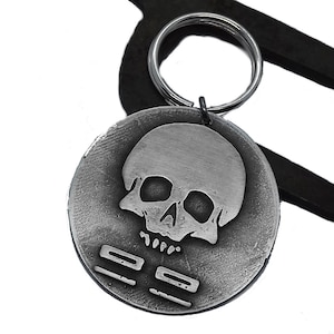 May include: A silver-colored metal keychain with a skull design. The round pendant features a skull in the center and two horizontal lines below. The keychain is attached to a metal ring. The background is white with dark metal shapes.