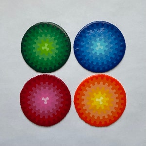 May include: Four circular coasters in vibrant colours. Each coaster has a gradient design with a gear-like pattern around the edge. The colours are green, blue, red, and orange, with lighter shades in the centre of each coaster.