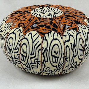 May include: A round, orange and white patterned pouf with a black and white graphic design. The pouf is made of fabric and has a tufted design.