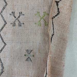 Op de afbeelding: Een beige kleed met zwarte en groene geometrische patronen. Het kleed heeft een rafelige rand en is geweven met een losse textuur.