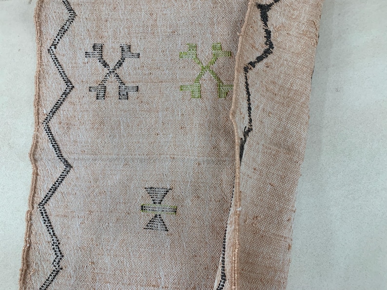 Op de afbeelding: Een beige kleed met zwarte en groene geometrische patronen. Het kleed heeft een rafelige rand en is geweven met een losse textuur.