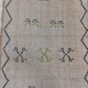 Op de afbeelding: Een beige kleed met een zwart en groen geometrisch patroon. Het patroon heeft herhalende kruisen en X-vormen.