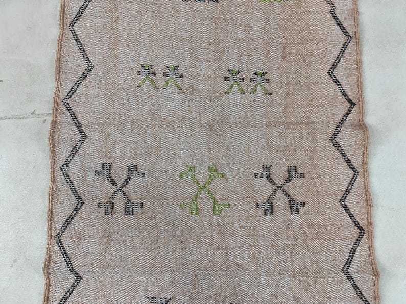 Op de afbeelding: Een beige kleed met een zwart en groen geometrisch patroon. Het patroon heeft herhalende kruisen en X-vormen.