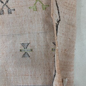 Op de afbeelding: Close-up van een beige geweven tapijt met zwarte en groene geometrische patronen. Het tapijt heeft een rafelige rand en is doormidden gevouwen.