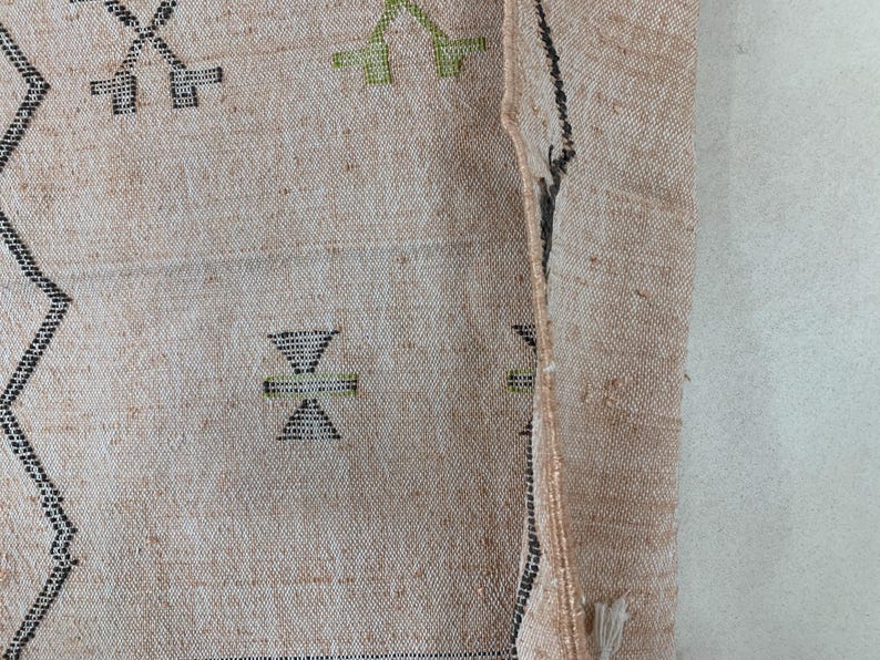 Op de afbeelding: Close-up van een beige geweven tapijt met zwarte en groene geometrische patronen. Het tapijt heeft een rafelige rand en is doormidden gevouwen.
