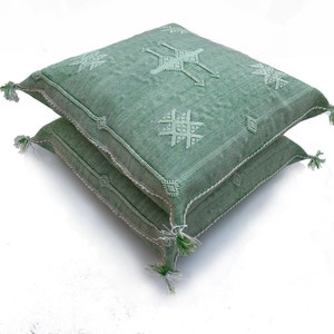 May include: Two green throw pillows with white embroidered geometric patterns. The pillows are stacked on top of each other.