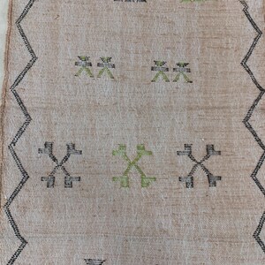Op de afbeelding: Een beige kleed met een zwart en groen geometrisch patroon. Het patroon heeft herhalende kruisen en X-vormen.