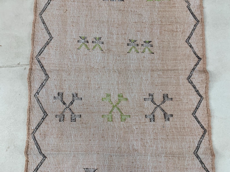 Op de afbeelding: Een beige kleed met een zwart en groen geometrisch patroon. Het patroon heeft herhalende kruisen en X-vormen.