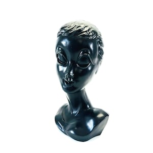 May include: A black mannequin head with a smooth, glossy finish. The head is tilted slightly to the side, and the eyes are closed. The mannequin is made of plastic and is designed to display jewelry or other accessories.