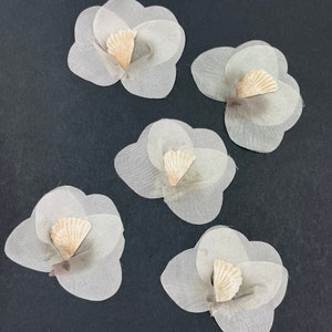 May include: Five white fabric flowers with a beige centre. The flowers are layered and have a delicate, sheer texture.