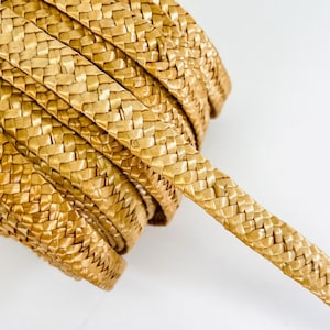 Può includere: Un rotolo di nastro di rafia intrecciata di colore marrone dorato naturale. Il nastro ha un aspetto piatto e intrecciato con una superficie strutturata. Il primo piano evidenzia l'intricata tessitura e la flessibilità del nastro, adatto per lavori artigianali e decorativi.